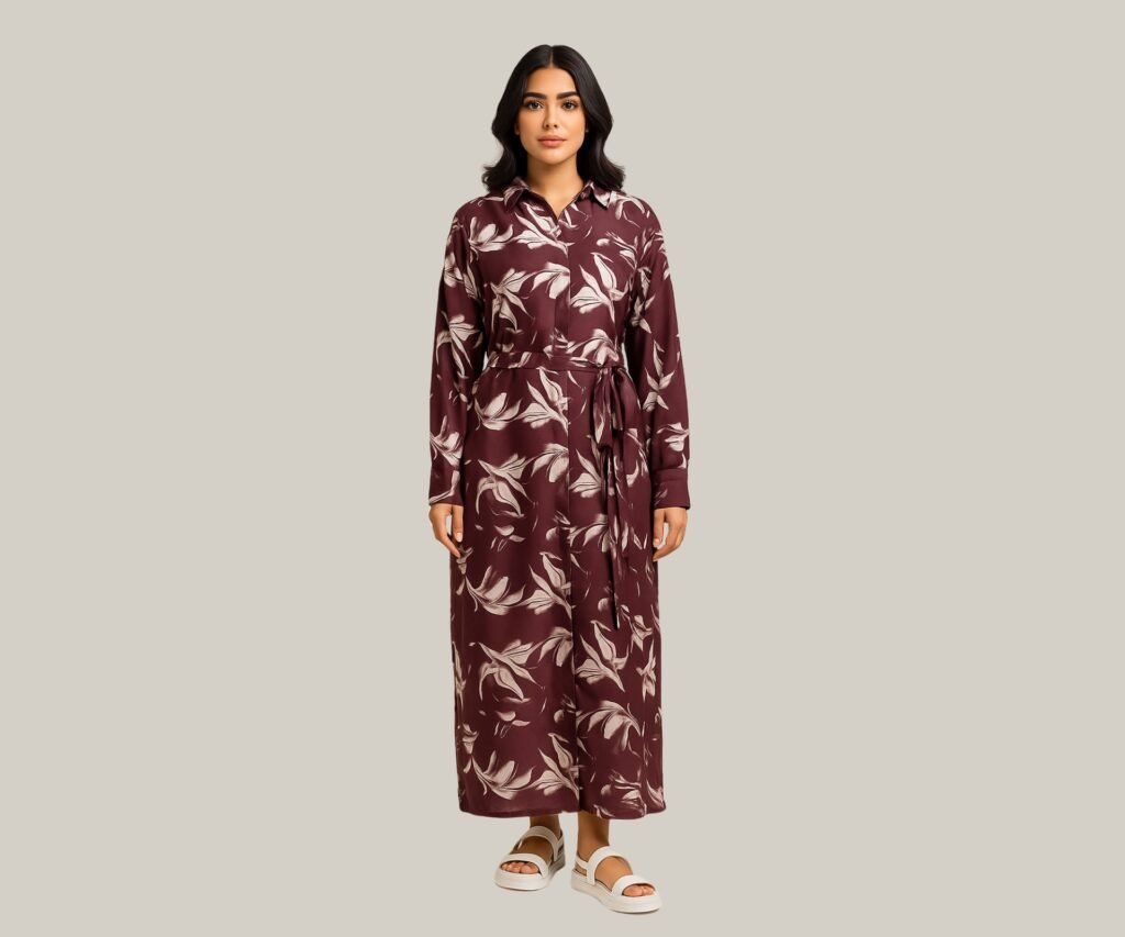Woman in a long, maroon floral Western dress with long sleeves and a tie waist, paired with white sandals, standing against a gray background.
